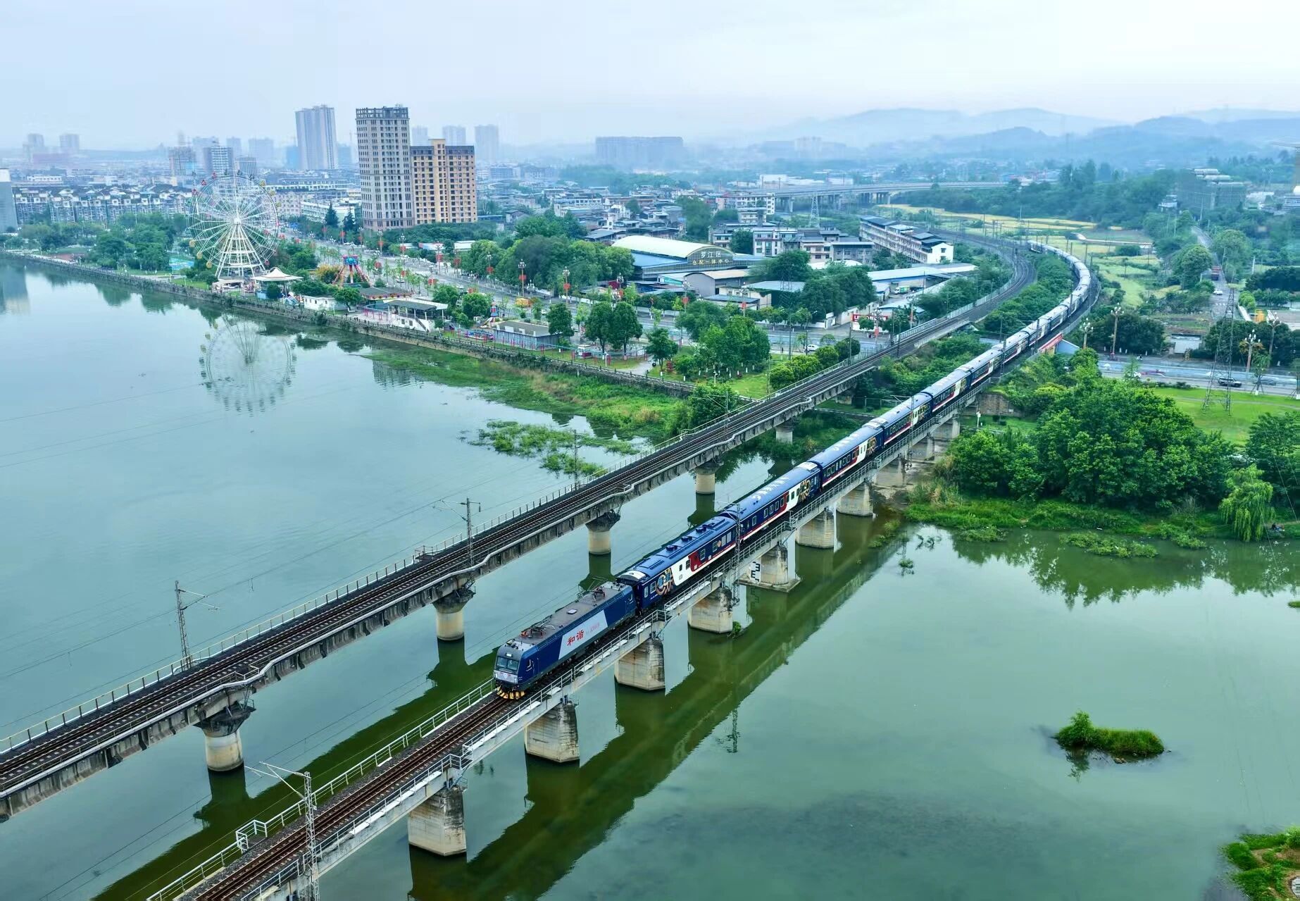 A luxury tourist train crossing a scenic viaduct over water, mountains and a distant city beyond.